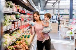 A woman with long brown hair holds a curly-haired baby on her hip while inspecting some packaged produce in the refrigerated section of a brightly lit grocery store.
