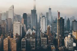 From a high angle, a six lanes of cars and taxis look very small in a dense metropolitan area with old buildings mingling with shiny new ones on a cloudy or smoggy day.