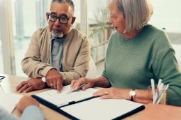 An elderly man and woman sit at a desk in a professional's office with a portfolio in front of them, discussing the file's contents.