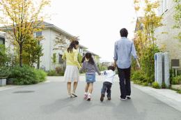 A man, woman, and two small children hold hands as they walk down a clean, bright neighborhood street with no cars and tidy trees and foliage.