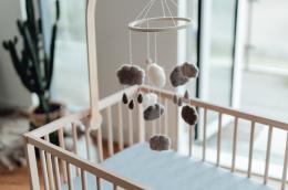 A baby's mobile featuring felted grey clouds and rain drops hangs over an empty wodden crib in a room with subdued natural light.