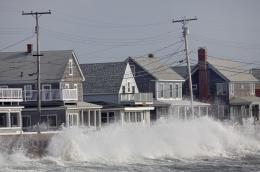 A turbulent ocean wave crashes against the seawall in front of a street of coastal homes on a cloudy day.