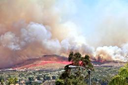 A large, active wildfire burns a hillside, approaching a shopping center, a residential neighborhood, and farmland while huge billows of smoke obscure the sky.