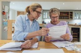 A middle-age and elderly woman sit in a sunny kitchen in front of a wooden table examining mail.