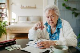 An elderly woman in a blue shirt and white cardigan sits in her apartment in front of bills, a ledger, and her wallet examining receipts.