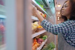 A woman in an apron and plaid shirt tidies up the refrigerated produce section in a grocery store.