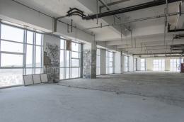 A large, empty interior space of a building under construction or renovation with unfinished concrete floors and exposed ceiling pipes and wiring.