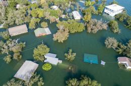 An aerial shot of a severely flooded neighborhood, where water has reached the roofs of several homes and the leafy tops of trees. Water has completely submerged several smaller structures, including a fenced-in backyard with patio furniture.
