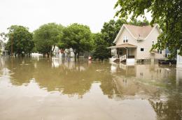 A severly flooded residential neighborhood with muddy water covering the streets and reaching up to the porches of houses, with trees, a stop sign, and electrical pole partially submerged as well.