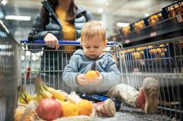 A toddler in a fuzzy blue sweater and red sneakers sits, holding and inspecting an orange, next to a couple bunches of bananas, a pomegranate, and a couple more oranges in a grocery cart pushed by a woman in a blue puffer jacket through an aisle of a grocery store lined on one side with crates of oranges.