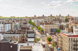 An elevated vantage point shows cars, busses, and people navigatinga street going through an urban setting dense with buildings and some trees.