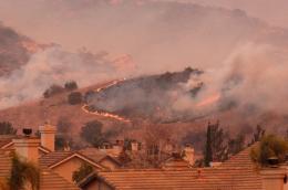 Smoke billows and a wildfire blazes on a hilltop adjacent to a neighborhood in an arid, hilly region.