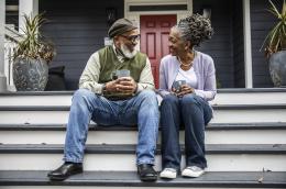 Two seniors sit on the steps in front of their home, holding mugs of coffee and smiling at each other..