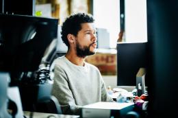 A young man with dark hair and a beard focuses on a computer screen in a communal working space.