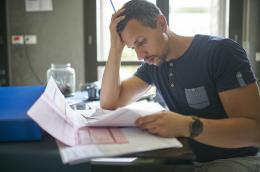 A man looks concerned as he looks through a stack of bills and other mail, with his head in his hand.
