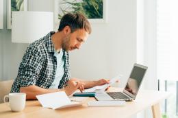 A man with light hair and a beard sits at a table in a brightly lit room in front of a laptop, scrutinizing one of a few sheets of paper and typing into his phone.