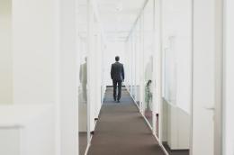 A man in a suit walks down a window-lined hallway in an office building