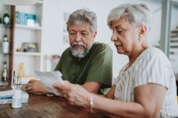 An elderly man and woman sit at their kitchen table analyzing a medical bill.