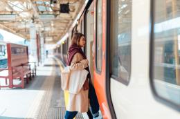 A young woman in a long, tan trench coat, blue jeans, and a dark red scarf steps aboard a public train from a covered platform.