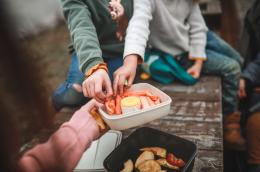 Two childen in sweaters reach for carrot sticks in a portable lunch container that's being held out by a third person, with whom they share a bench outside.