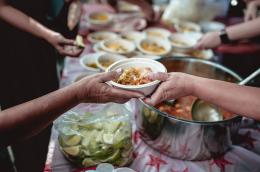A bowl of soup, ladled from a large, metal pot sitting next to a plastic bag of limes, is handed from one person to another.