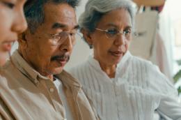 An elderly man and woman inspect something off camera, maybe something on a desk, along with a younger woman who appears to be explaining or helping.