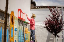 A muralist in a red shirt and straw hat stands on a short wooden ladder, holding a palette in one hand and a paintbrush in the other, adding on to a mural of colorful homes. 