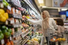 A woman pushing a grocery cart down a store's produce aisle reaches for an apple, with her little girl seated at the front of the cart and some oranges in the basket.