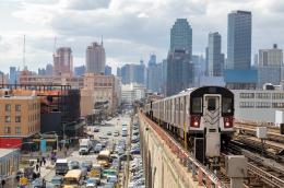 A busy urban skyline on a cloudy day, with lots  of people walking and cars visible on the street and parked, with a public train heading toward the camera.