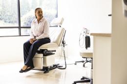 An anxious-looking woman in blue slacks and a pale pink button-down shirt sits with her hands clasped together on an exam chair in a doctor's office.