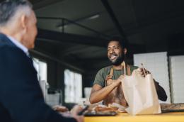 A man in a green t-shirt and a work apron covered in flour places a loaf of bread in a brown paper bag for a customer.