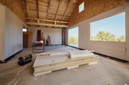 Tools and wooden panels sit on the ground of a home under construction on a sunny day, and other construction materials line the far wall under exposed beams.