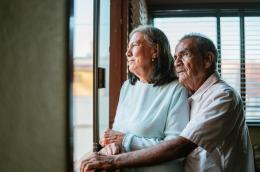 An elderly man embraces an elderly woman as they look out a window, presumably in their home.
