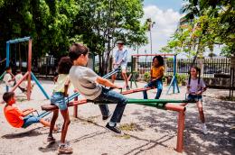 A group of young children play on the swings and three seesaws in a sunny playground. 