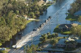An aerial shot of a deeply flooded neighborhood, with a line of cars fording the flooded streets and homes looking like islands surrounded by water. 