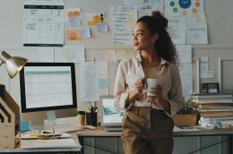 A young woman in a business-casual outfit holds a coffee mug, looking out a window to her right, standing in front of her desk, which features plenty of evidence of productivity: a calendar, a stack of books, motivational quotes, a desk top, a laptop, and even a backup cup of coffee.