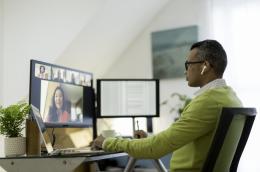 A man wearing a business casual shirt and sweater sits in front of a desk with a laptop and two additional monitors, holding a virtual meeting with colleagues.