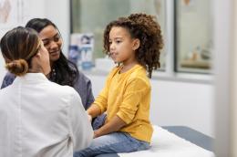 A young child of color sits on the examiniation table in a doctor's office as a female doctor takes her vitals while her mother smiles at her daughter.