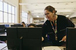 An elder Indigenous woman wearing beaded earrings and a colorful lanyard with many keys and , helps a person working at a computer in a shared space, perhaps a library.