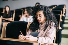 A young woman with curly hair, cat-eye glasses, and a light pink hoodie sits among a few other students in a classroom, writing in a notebook with a pencil.