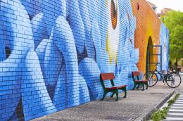 A colorful blue and orange mural covers the brick wall running alongside a street, with a couple benches and a bike rack.