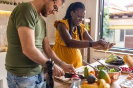 A father chops a tomato into quarters as a pregnant woman squeezes lemon over a salad they're preparing together at their kitchen island, which also holds bowls of different kinds of fruits, veggies, and a carton of eggs.