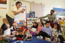 A class of small children sit in a circle in a classroom as the teacher in a yellow shirt and glasses presents on a strawberry, with the teacher's aid sittings nearby, looking at a diagram of the solar system.