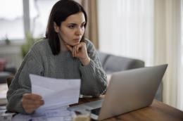 A woman with short brown hair and a sage-colored cable sweater sits at a countertop, holding a bill with one hand while supporting her chin with the other and looking at her laptop with a furrowed brow.