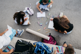 A group of children sit outdoors on pavement in a circle, drawing in notebooks with colorful pencils and chalk scattered around them. Backpacks and art supplies lie nearby, and one child holds a piece of paper while another eats an apple.
